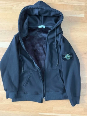 Svart kofta från Stone Island - Säljer en svart kofta från Stone Island med dragkedja, huva med dragsko och mjukt foder. Klassisk patch med logga på vänster ärm och två fickor framtill. Perfekt för dig som gillar streetwear och vill ha något stilrent och bekvämt.