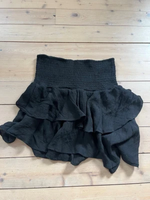 Svart volangkjol  - Säljer en svart kortkjol med bred smock i midjan och flera lager volanger. Kjolen har en luftig och flowy känsla, perfekt för dig som gillar en trendig och bekväm stil. Materialet känns lätt och mjukt mot huden. Aldrig använd men lappen är bortklippt.