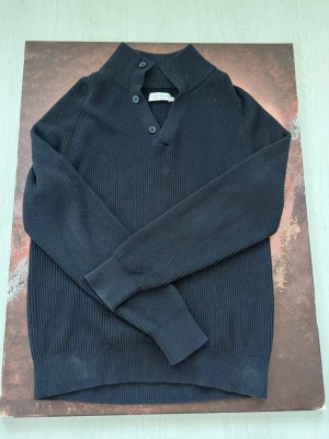 Mörkblå stickad tröja Casual friday  - Snygg mörkblå stickad tröja från Casual friday med ribbad struktur och tre knappar vid halsringningen. Tröjan har lång ärm och breda muddar vid ärmslut och nederkant. Perfekt för dig som gillar stilrena och tidlösa plagg.Tröjan är fint skick utan defekter och är i storlek XL!