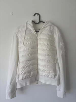 Vit dunväst med huva från Moncler - Snygg vit dunväst från Moncler med huva och dragkedja framtill. Västen har quiltad framsida, släta ärmar och fickor med dragkedja. Moncler-logga på ärmen och sportig design. Perfekt för lager-på-lager och streetstyle.