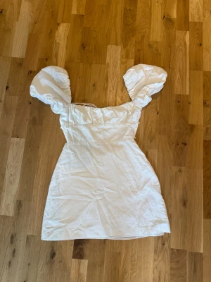 Vit kort klänning med puffärmar - Supersöt vit kort klänning med puffiga ärmar och snörning i ryggen. Klänningen har en feminin siluett och volangdetaljer vid bysten. Perfekt för sommarens alla tillfällen och ger en romantisk vibe.