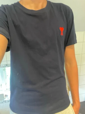 Mörkblå t-shirt från Ami Paris - Mörkblå t-shirt från Ami Paris med rakt snitt och rund hals. På bröstet finns Ami:s ikoniska röda hjärta med bokstaven A broderat. Tillverkad i mjuk bomull och har korta ärmar. Perfekt för en clean och stilren look.