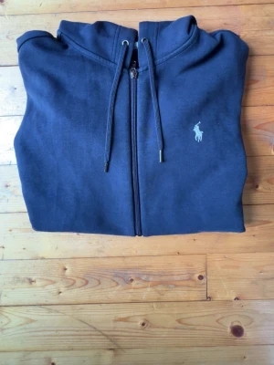 Mörkblå hoodie från Polo Ralph Lauren - Mörkblå hoodie med dragkedja från Polo Ralph Lauren. Klassisk design med huva och dragsnören, två fickor framtill och den ikoniska broderade loggan på bröstet. Tillverkad i mjuk bomullsblandning för en skön och avslappnad stil.