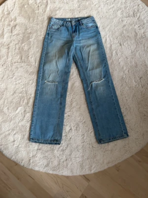 Ljusblå raka jeans - Säljer ett par ljusblå jeans med raka ben och slitningar vid knäna. Jeansen har klassisk femficksdesign och är tillverkade i denim.