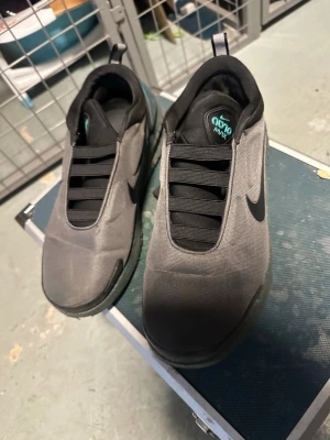 Nike Go FlyEase grå sneakers - Säljer ett par Nike Go FlyEase sneakers i grått med svart swoosh och elastiska band istället för vanliga skosnören. Skorna har en futuristisk design och är tillverkade i textil med detaljer i syntet. Sulan är tjock och har en cool mintgrön detalj på hälen.