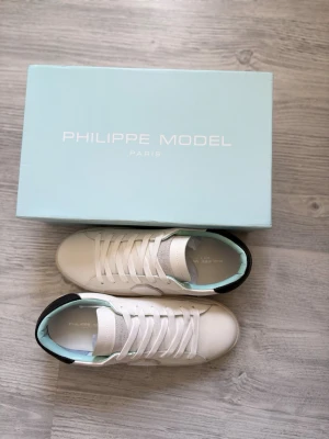 Vita Philippe Model sneakers - Snygga vita sneakers från Philippe Model med klassisk låg siluett. Skorna har vita skosnören, svart detalj på hälen och diskret logotyp på sidan. Tillverkade i läder med ljusblå insida för en fräsch look. Perfekta för en stilren och trendig outfit.