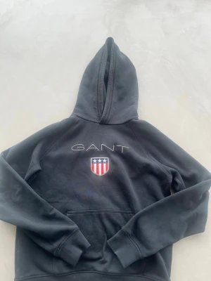 Svart hoodie från GANT med logga - Säljer en svart hoodie från GANT med stor huva och känguruficka. På bröstet finns GANT-loggan och ett broderat emblem med stjärnor och ränder i rött, vitt och blått. Hoodien har ribbade muddar vid ärmar och nederkant. Perfekt för en avslappnad och stilren look.
