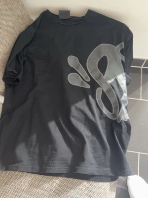 Syna world T-shirt och Shorts - Säljer en svart t-shirt med stor, grå graffiti-inspirerad print över både fram- och baksida. T-shirten har klassisk passform och korta ärmar. Perfekt för dig som gillar streetwear och vill sticka ut med en unik design. Passar L/M med  