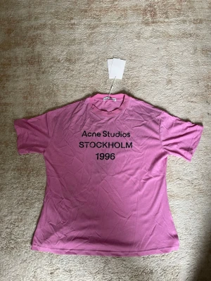 Rosa Acne Studios t-shirt med tryck - Snygg rosa t-shirt från Acne Studios med svart texttryck 'STOCKHOLM 1996' på bröstet. Klassisk passform med rund hals och korta ärmar. Perfekt för en trendig och avslappnad stil.
