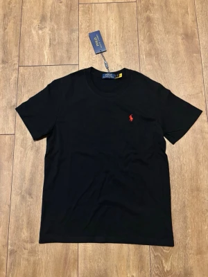 Helt ny Polo Ralph Lauren T-shirt – Svart med röd logga - Säljer en helt ny och oanvänd t-shirt från Polo Ralph Lauren i svart med en snygg röd logotyp, vilket syns i bilderna. Originaletikett sitter kvar. Passar perfekt för en stilren och klassisk look. Storlek: S