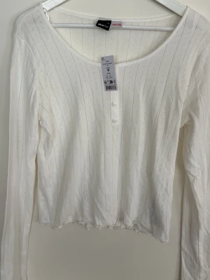 Vit ribbad långärmad topp Gina Tricot - Snygg vit långärmad topp från Gina Tricot med ribbad struktur och knappar framtill. Toppen har en rund halsringning och lätt croppad passform med vågig kant nedtill. Perfekt att styla med jeans eller kjol.