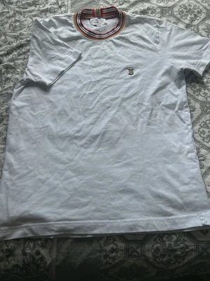 Vit Burberry t-shirt med randig krage - Snygg vit t-shirt från Burberry med klassisk randig krage i rött, svart, vitt och beige. T-shirten har korta ärmar och diskret Burberry-logga broderad på bröstet. Tillverkad i mjuk bomull för en skön känsla.