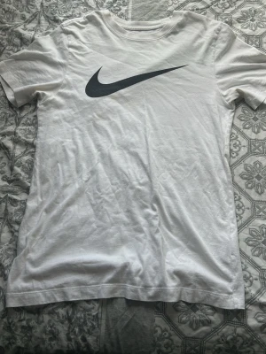 Vit Nike t-shirt med stor logga - Klassisk vit t-shirt från Nike med stor svart swoosh-logga på bröstet. Tillverkad i mjuk bomull och har en normal passform med korta ärmar och rund hals. Perfekt för en sportig och avslappnad stil.