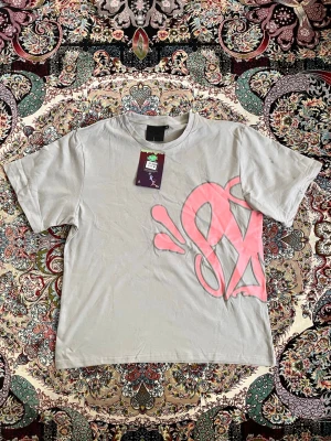 Grå t-shirt med rosa tryck - Säljer en grå t-shirt med ett stort rosa grafiskt tryck på framsidan. T-shirten har klassisk passform och korta ärmar. Materialet är mjuk bomull som känns skön mot huden. Perfekt för dig som gillar streetwear och vill sticka ut lite extra.