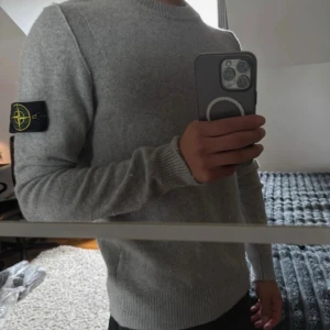 Grå stickad tröja från Stone Island - Snygg grå stickad tröja från Stone Island med klassisk rund halsringning och ribbade muddar. Tröjan har den ikoniska loggan på vänster ärm och är tillverkad i ett mjukt material som passar perfekt till höst och vinter.