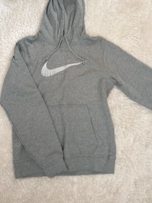Grå Nike hoodie med logga - Snygg grå hoodie från Nike med stor vit logga på bröstet i ett mönstrat tryck. Hoodien har huva med dragsko och en klassisk magficka. Tillverkad i mjukt sweatshirtmaterial som är perfekt för chill dagar.