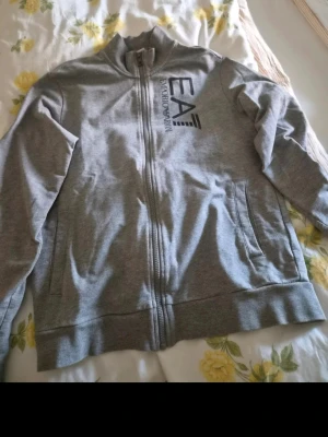 Grå zip-hoodie från Emporio Armani - Snygg grå hoodie från Emporio Armani med dragkedja framtill och stort EA7-tryck på bröstet. Tröjan har ståkrage, långa ärmar och två fickor på sidorna. Perfekt för en avslappnad och trendig stil.