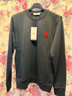 Svart sweatshirt från Ami Paris - Stilren svart sweatshirt från Ami Paris med klassisk rund halsringning och röd Ami-dekal broderad på bröstet. Tröjan har långa ärmar, ribbade muddar och är tillverkad i mjuk bomull för maximal komfort.