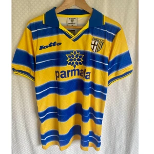Vintage Jersey AC Parma Calcio 1999 - Klassisk Parma AC fotboll hemmatröja säsong 1998/1999 från Lotto med CRESPO 9 på ryggen. Tröjan är gul med breda blå horisontella ränder, blå krage och detaljer. Parmalat-logga på bröstet och klubbmärke. Tillverkad i lätt polyester, perfekt för dig som älskar retrostil.