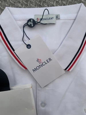 Vit Moncler pikéskjorta med ränder - Snygg vit pikéskjorta från Moncler med klassisk krage och knappar framtill. Kragen har stilrena röda och svarta ränder och bröstet pryds av Monclers ikoniska logotyp. Perfekt för dig som gillar exklusiva och tidlösa plagg.