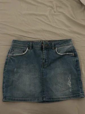 Blå jeanskjol från ONLY - Snygg blå jeanskjol från ONLY med klassisk femficksdesign, slitna detaljer och ljus tvätt. Kjolen har bälteshällor, knapp- och dragkedjestängning samt råa kanter vid fickorna. Perfekt för en avslappnad och trendig look.