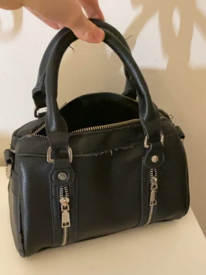 Svart handväska från Misako - Snygg svart handväska från Misako i konstläder med två korta handtag och silverfärgade dragkedjor framtill. Väskan har en boxig form och detaljer i metall som ger en cool look. Perfekt för dig som gillar stilrena accessoarer med lite edge.