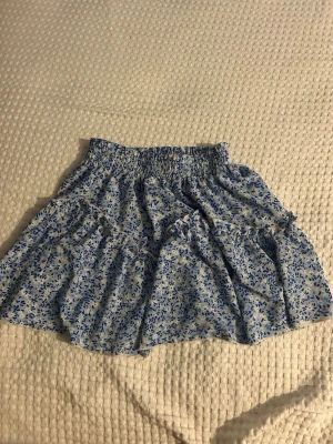 Blommig volangkjol från SHEIN - Söt kort kjol från SHEIN med blått blommönster på vit botten. Kjolen har volangdetaljer och resår i midjan för en bekväm passform. Perfekt för en fräsch och somrig look.