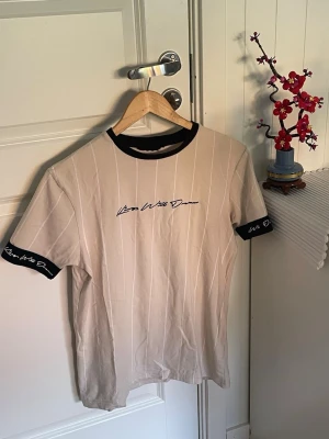 Beige med vita ränder t-shirt från Kings Will Dream - Snygg beige t-shirt från Kings Will Dream med tunna vertikala ränder och svart text ”Kings Will Dream” på bröstet. Svart ribbad krage och svarta muddar med logga på ärmarna ger en clean kontrast. Perfekt till jeans eller shorts. Etiketten klipptes bort pga irritation från den när man tog på sig tröjan, men storlek minns som S, annars använd dig av måtten på bilderna