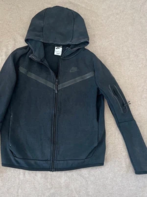 Svart Nike hoodie med dragkedja - Snygg svart hoodie från Nike med dragkedja framtill och huva. Har en diskret Nike-logga på bröstet och en cool detalj med ficka på ena ärmen. Tillverkad i mjukt material och har en sportig look med svarta ränder över bröstet.