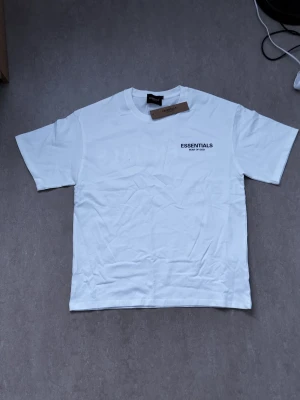 Vit Essentials Fear of God t-shirt - Säljer en vit t-shirt från Fear of God Essentials med svart logga tryckt på bröstet och stor text på ryggen. Klassisk passform och rund hals. Perfekt basic-plagg för dig som gillar streetwear och enkel stil.