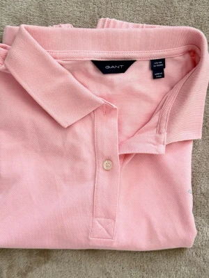 Ljusrosa pikétröja från GANT - Säljer en ljusrosa pikétröja från GANT med klassisk krage och knappslå framtill. Tröjan är tillverkad i mjuk bomull och har en stilren look som passar perfekt till jeans eller kjol. Enkel och snygg design med diskret logga på ärmen.