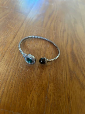 Silverfärgat armband med grön sten - Snyggt armband i silverfärg med tvinnad design. Ena änden har en stor rund grön sten omgiven av små klara stenar, och andra änden har en svart pärla med dekorativa detaljer. Perfekt accessoar för att lyfta din stil.