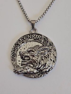 Silverfärgat Fenrir-halsband - Säljer ett silverfärgat halsband med en rund medaljong som har en detaljerad relief av Fenrir, den mytologiska vargen, och texten 'FENRIR' i runliknande stil. Hänget har vikingainspirerade mönster och hänger på en kedja i metall. Perfekt för dig som gillar nordisk mytologi och unika smycken. Materialet rostfritt stål 