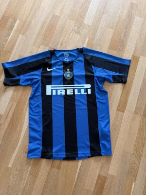 Inter Milan matchtröja Nike blå/svart - Säljer en klassisk Inter Milan matchtröja från Nike med blå och svarta vertikala ränder. Tröjan har Pirelli-logga på bröstet, klubbmärke och Nike swoosh. Tillverkad i lätt och ventilerande material, perfekt för fotboll eller streetwear.