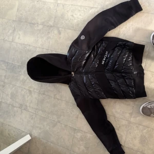 Svart cardigan från Moncler - Svart cardigan från Moncler med glansiga partier på bröst och rygg, samt Moncler-logga på ärmen. Jackan har huva, dragkedja framtill och är långärmad. Perfekt för kyligare dagar och har en modern, sportig look.