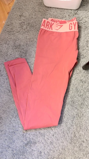 Rosa leggings från Gymshark - Snygga rosa leggings från Gymshark med hög midja och bred resår med logga. Tillverkade i ett stretchigt och mjukt material som sitter skönt på kroppen. Perfekta för träning eller chill. Klassisk Gymshark-design med diskret logotyp på benet.