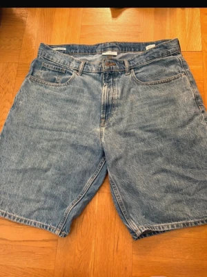 Blå jeansshorts  - Klassiska blå jeansshorts med fem fickor och baggy passform. Shortsen har en midwaist design och är tillverkade i slitstark bomull. Perfekta för sommaren och har en tidlös look med knapp och dragkedja framtill.
