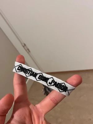 Christian Dior Junior armband - Snyggt armband från Christian Dior Junior i vitt tyg med svart broderad text och grafiskt mönster. Armbandet har tofsar i ändarna och en cool, ungdomlig vibe. Perfekt accessoar för att lyfta din outfit.
