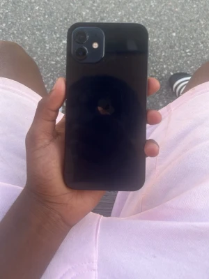Svart iPhone 12 med dubbla kameror - Säljer en svart iPhone 12 med stilren design och rundade kanter. Baksidan är i glas och mobilen har två kameror i hörnet. Klassisk Apple-logga på baksidan och en snygg, modern look som passar perfekt för dig som gillar teknik.