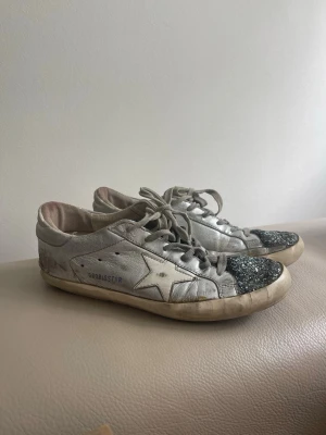 Golden Goose superstar sneakers  - Säljer ett par ikoniska silvriga sneakers från Golden Goose i silverfärgat läder med glittriga svarta tår och den klassiska stjärnan på sidan. Skorna har vita snören och en vintage-inspirerad sula. Perfekta för dig som gillar en unik och trendig look. Box följer med. Storlek 40 italiensk vilket motsvarar 39 svensk. 