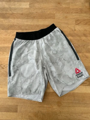 Reebok Crossfit Games shorts grå - Snygga gråa Reebok Crossfit Games shorts med marmorerat mönster och svart midjeresår. Shorts med två svarta dragkedjefickor och tryck fram och bak. Perfekta för träning och sommarens alla aktiviteter.