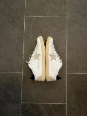 Golden Goose vita sneakers med stjärna - Golden Goose sneakers i vitt läder med grå stjärna på sidan och beige sula. Klassisk låg modell med svarta detaljer vid hälen och grå skosnören. Medföljer original dustbag med tryck. Perfekta för dig som gillar en avslappnad och trendig stil.