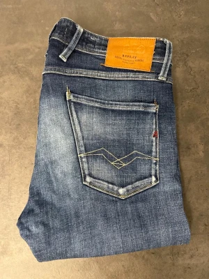 Blå jeans från Replay, stl 33/34 - Snygga blå jeans från Replay med klassisk femficksdesign och smal passform. Jeansen har slitningar och ljusare partier för en trendig look samt detaljerad söm på bakfickan. Tillverkade i denim med skön stretch.