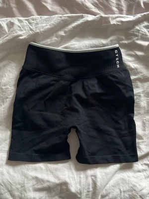 Svarta träningstights från Gtech - Säljer ett par svarta träningstights/shorts från Gtech med hög midja och bred resår. Materialet är stretchigt och följsamt, perfekt för gym eller löpning. Diskret logga på linningen och sömlös design för extra komfort.