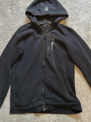 Svart hoodie från Sail Racing - Svart hoodie från Sail Racing med dragkedja framtill och en bröstficka med logga. Tillverkad i mjukt material med fleece på insidan för extra komfort. Huva med broderad detalj och långa ärmar. Perfekt för en avslappnad och sportig stil.