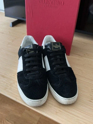 Valentino Garavani svarta sneakers - Snygga sneakers från Valentino Garavani i svart mocka med vita detaljer och vit sula. Klassisk låg modell med svart snörning och logga på plösen. Perfekta för dig som vill ha en stilren och exklusiv look.