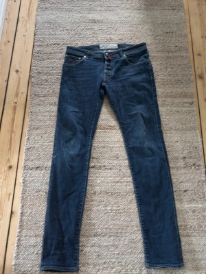 Slim fit Jacob cohen - Säljer ett par blå skinny jeans från 7 For All Mankind med klassisk femficksmodell och röda detaljer på bakfickorna. Jeansen har normal midja och är tillverkade i mjukt denimtyg som sitter snyggt och bekvämt.