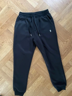 Svarta mjukisbyxor från Ralph Lauren - Säljer ett par svarta mjukisbyxor från Ralph Lauren med dragsko i midjan och resår vid bensluten. Byxorna har två sidofickor, en bakficka och den klassiska vita Polo-loggan broderad på ena benet. Perfekta för chill dagar eller när du vill vara bekväm.