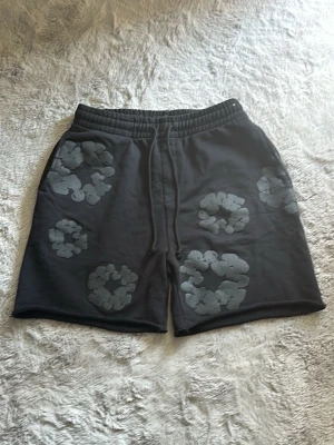 Svarta shorts från Denim Tears - Svarta shorts från Denim Tears med unika gråa blommönster över hela plagget. Shortsen har resårmidja med snörning för skön passform och är tillverkade i mjuk bomull. Perfekta för en chill och avslappnad stil.