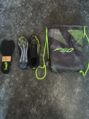 Adidas F50 fotbollsskor svart/neon - Adidas F50 fotbollsskor i svart med neon-gula detaljer och mönster. Skorna har strömlinjeformad design, plastdobbar och medföljer innersulor samt en F50 påse. Perfekta för spel på gräsplan, tillverkade i syntetmaterial för lätt vikt och snabbhet.